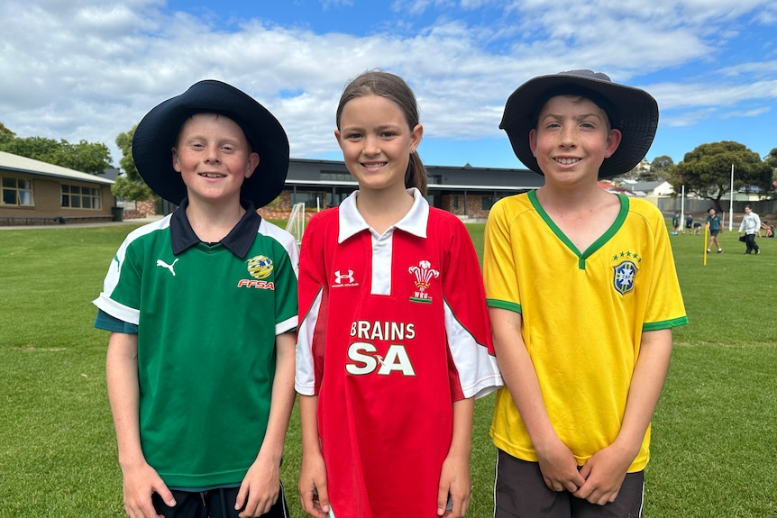 Young kids, a boy on the left in a green jersey, a girl in a red jersey and a boy on the right in yellow.