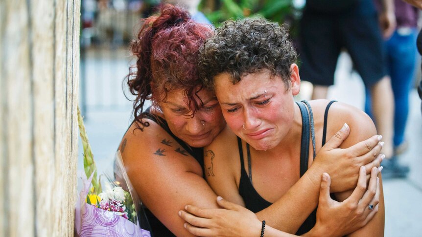 A mother hugs her crying daughter as both sit next to flower bouquet