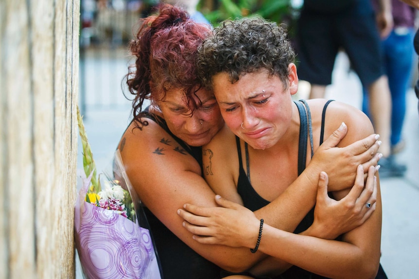 A mother hugs her crying daughter as both sit next to flower bouquet