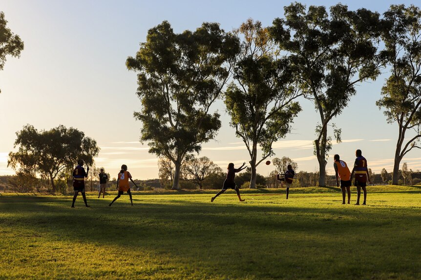 Aboriginal women wearing team uniforms kick the football in the late afternoon sun on a green football ground amid trees