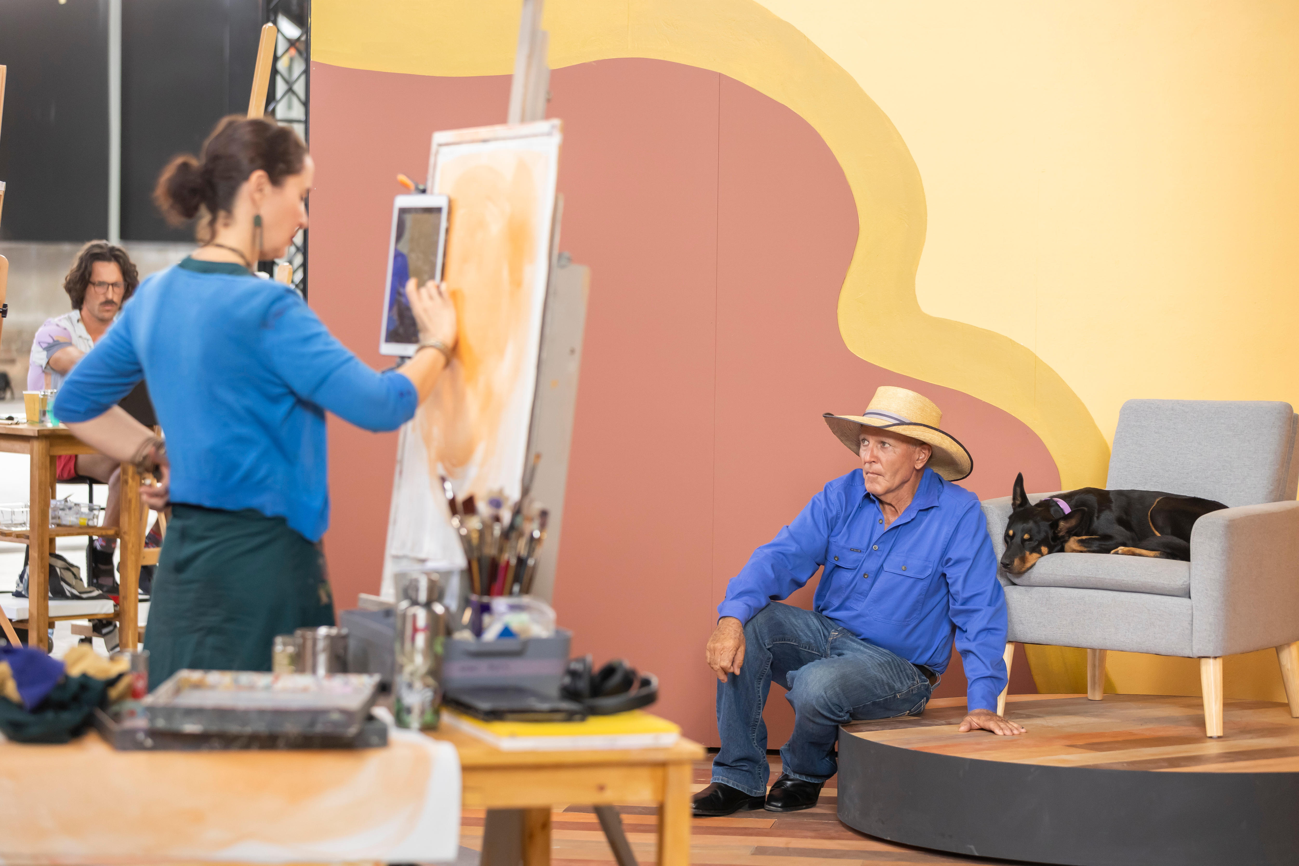 A woman painting a portrait of a man and dog on a TV set.