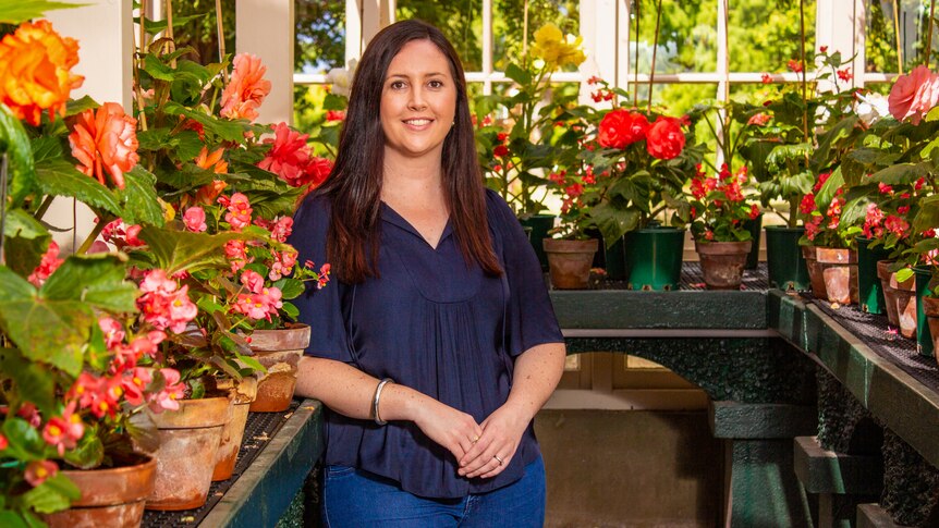 A woman with long dark hair, wearing a navy top, standing in a conservatory with potted flowers in the background.