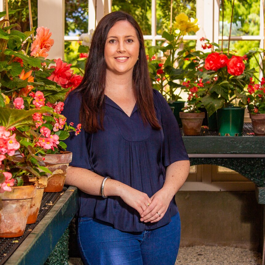 A woman with long dark hair, wearing a navy top, standing in a conservatory with potted flowers in the background.