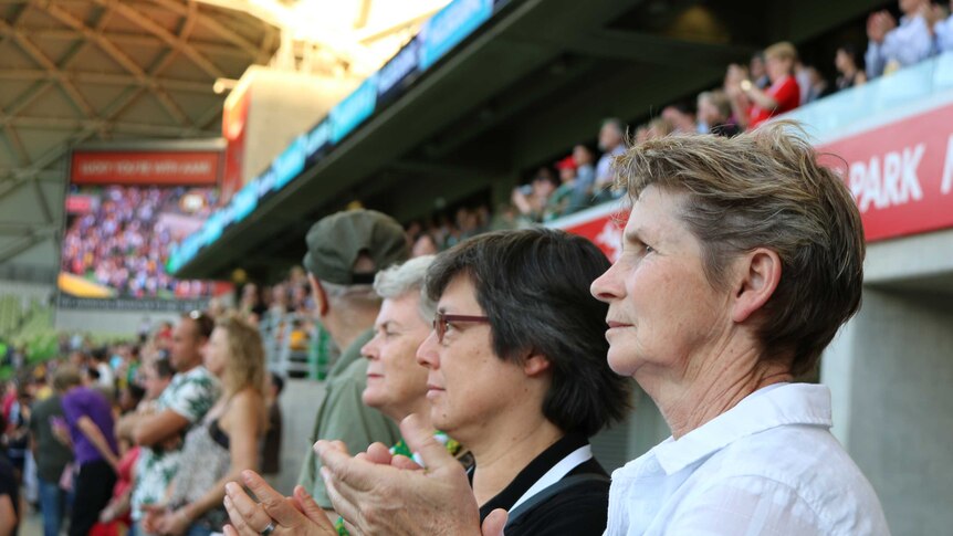 Three women at a football game stand in the crowd, clapping