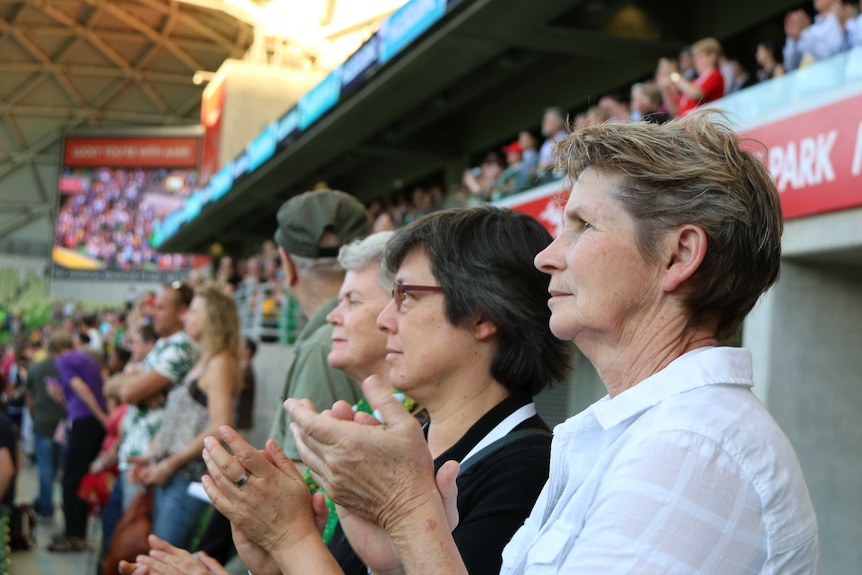 Three women at a football game stand in the crowd, clapping