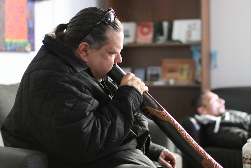 Man in black coat indoors playing a didgeridoo with another man in background lying on couch