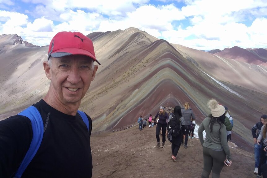 A selfie taken by a man in a red cap in front of a mountain with other tourists in the background.