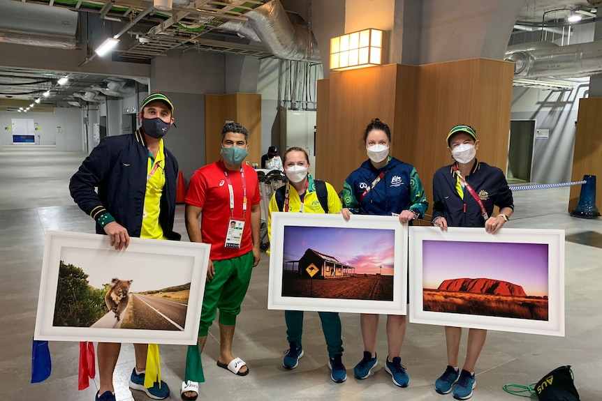 A middle eastern man in red and green stands next to four caucasian people who hold paintings of Australian scenes.