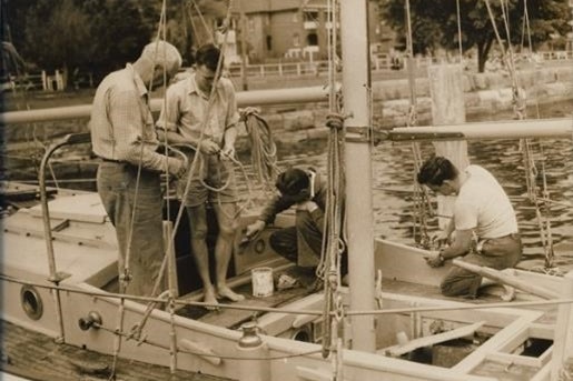 An older man stands on board a boat showing a younger man how to knot a rope, while two other sailors crouch down.