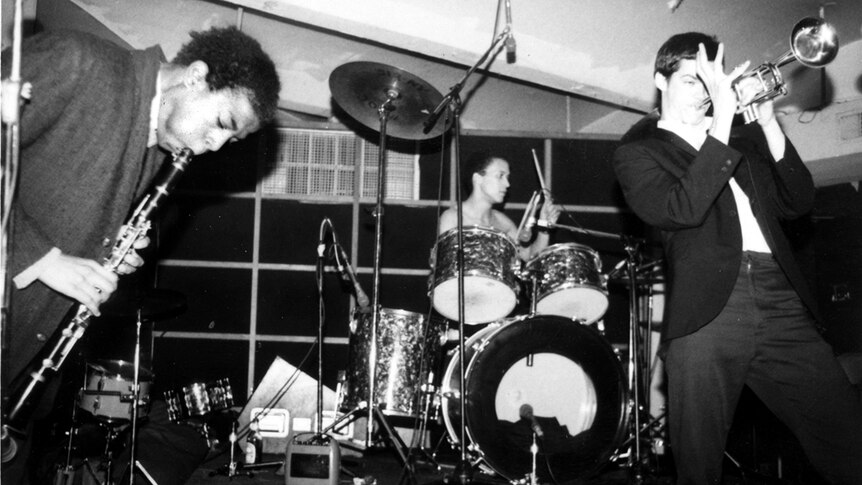 Black and white photo of Jean-Michel Basquiat performing clarinet with Michael Holman on drums and Shannon Dawson on trumpet.