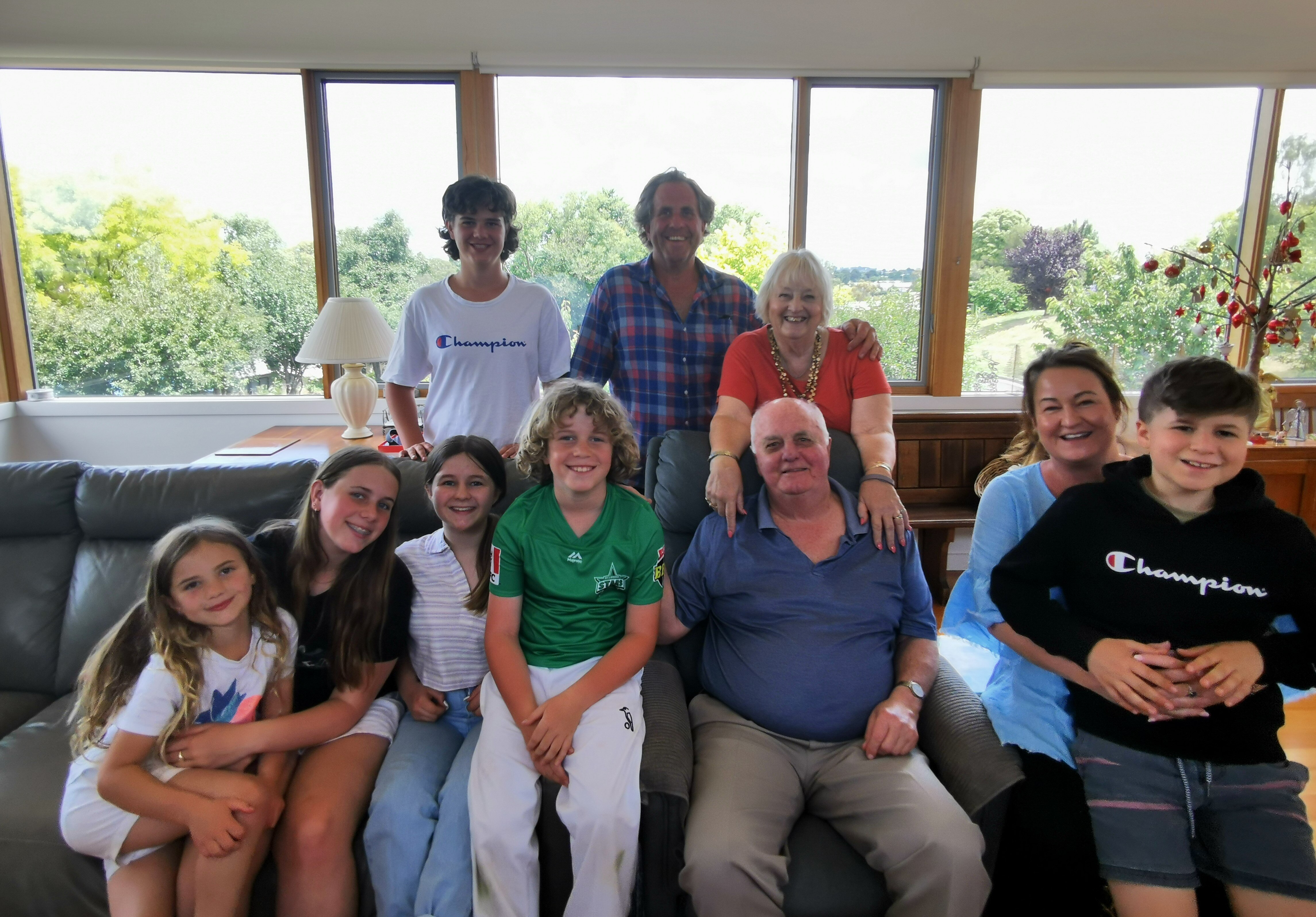 An older man sits on a couch surrounded by his wife and members of his extended family