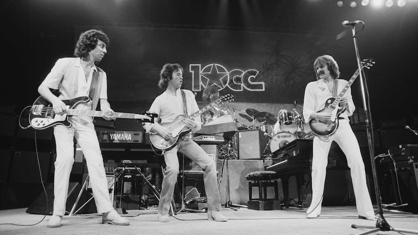 A black and white photo of rock musicians on stage with guitars. The band's name 10cc is written at the back of the stage.
