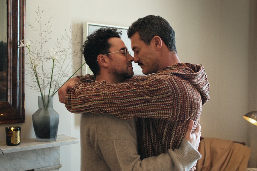 A film still of Daniel Levy and Luke Evans, embracing, their faces almost touching, in a living room.