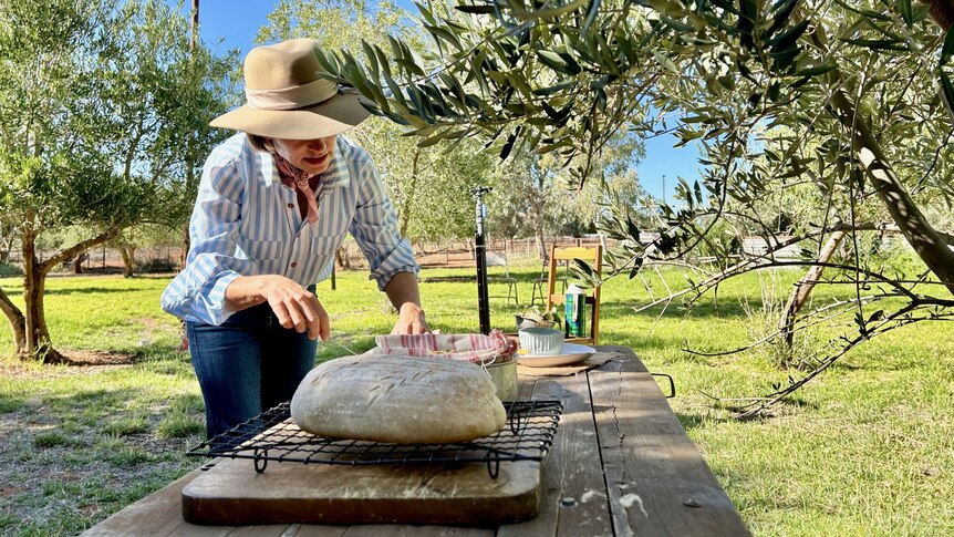 A loaf of bread on a table and a women preparing the table with baked goods amongst olive trees in sheep yards