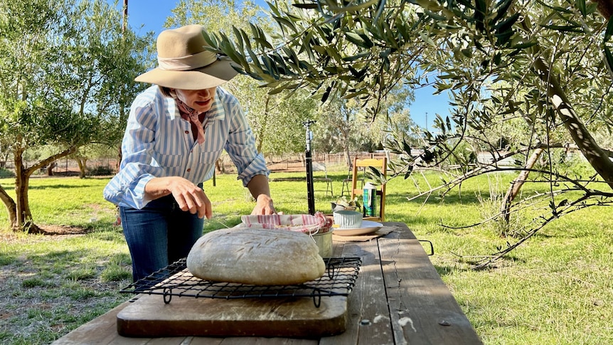 A loaf of bread on a table and a women preparing the table with baked goods amongst olive trees in sheep yards