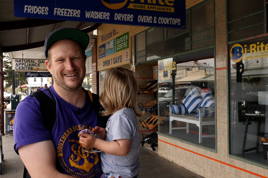 Smiling, man of 40 years old holding toddler out front of hardware store in Port Fairy wearing purple t-shirt.