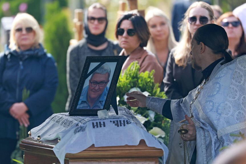 A crowd gather around a casket with a framed photo of a man on it. 
