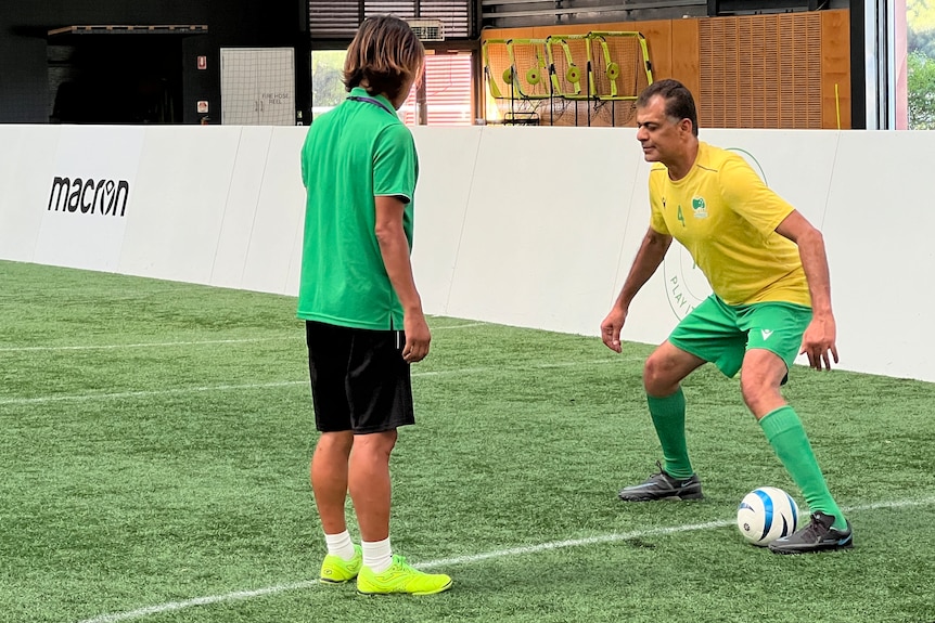 A male blind footballer has his knees bent and is placed over the ball while a coach watches on to assist.