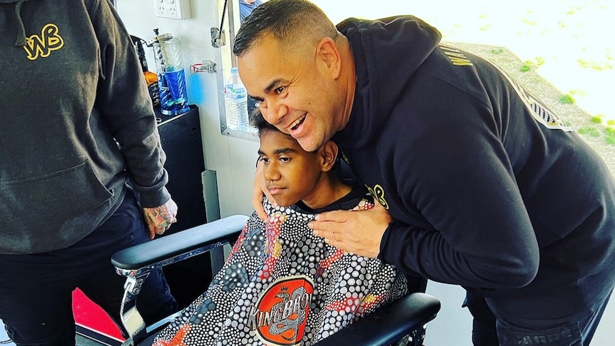 a young boy sits in a barbers chair with the barber holding him smiling in a black hoodie.
