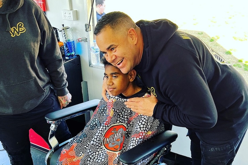 a young boy sits in a barbers chair with the barber holding him smiling in a black hoodie.