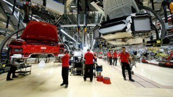 Men in red shirts work on a production line with a red Porsche hanging from the roof.