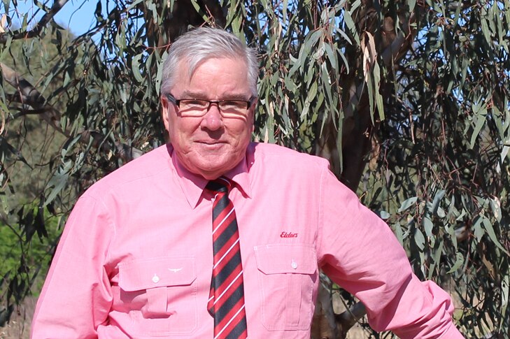Older man with neat, grey hair and black glasses wearing a pink shirt and black and red tie standing by a farm fence.