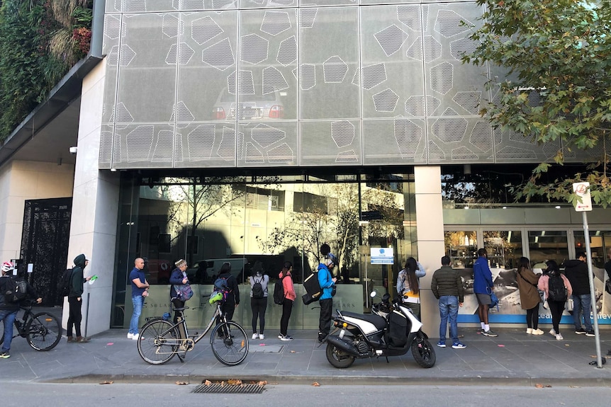 Fourteen people stand in a line outside a city building, waiting to collect meals.