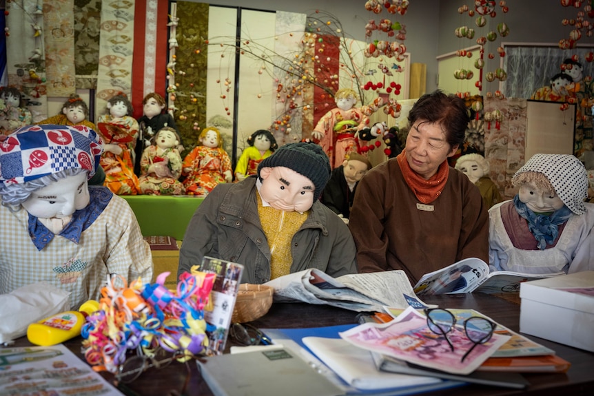 A woman wearing a red scarf and jumper looks at a doll that is hunched over a paper.