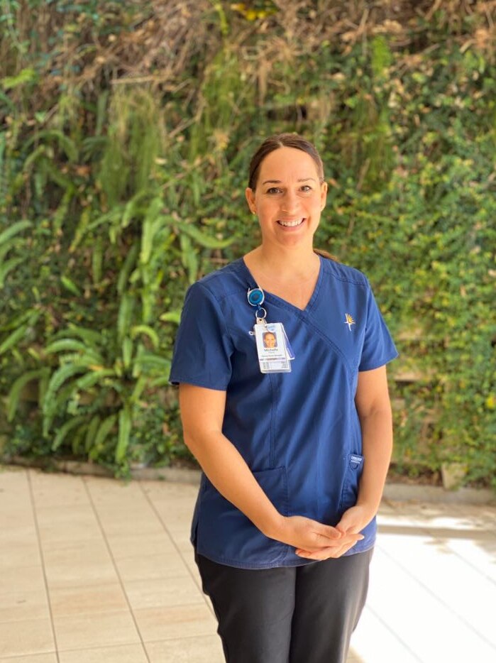A nurse with dark hair and blue nurse uniform standing in front of a leafy wall