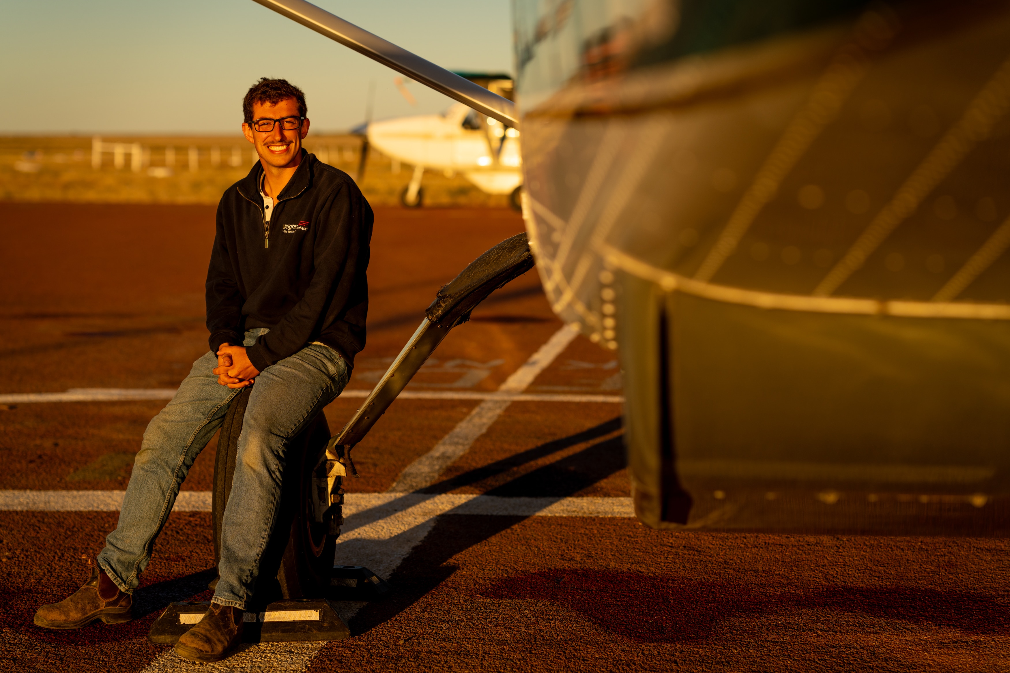 A young man sitting on the tire of a small airplane, smiling at the camera