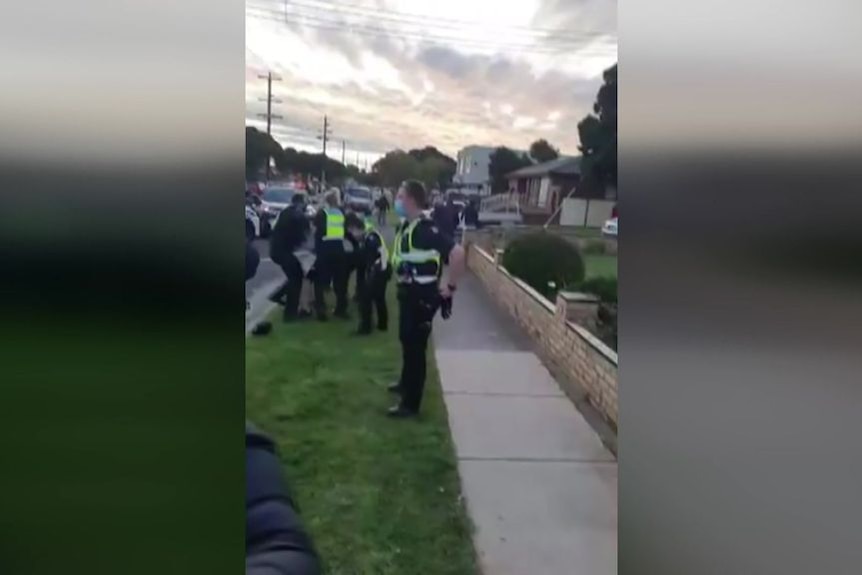 Police speak to people walking along a suburban street while other officers arrest a man in the background.
