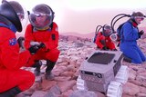 Students and man in orange and blue space suits collect rocks in rocky terrain simulated crater beside rover