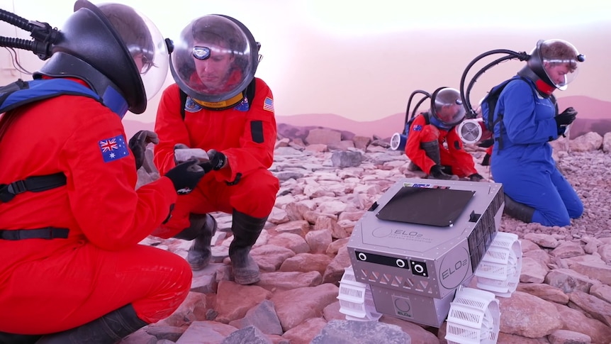 Students and man in orange and blue space suits collect rocks in rocky terrain simulated crater beside rover
