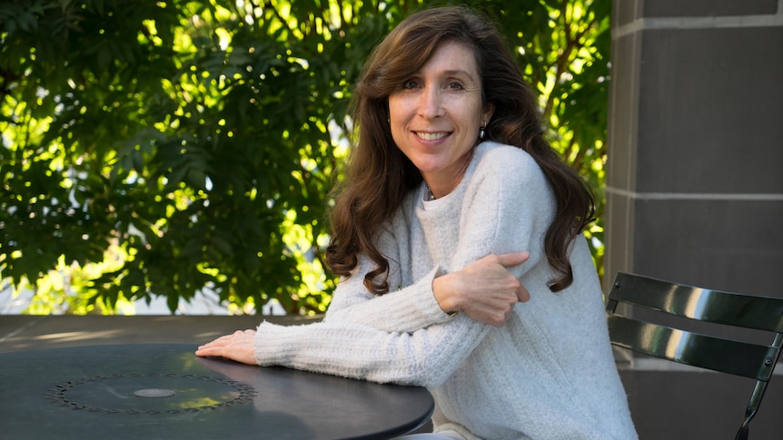 Middle-aged woman with long brown hair sits at a table and smiles. 