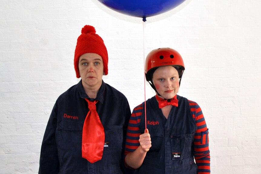 Two white women in motley navy and red suits and clown makeup. One wears a red beanie, the other a helmet and holds balloon.