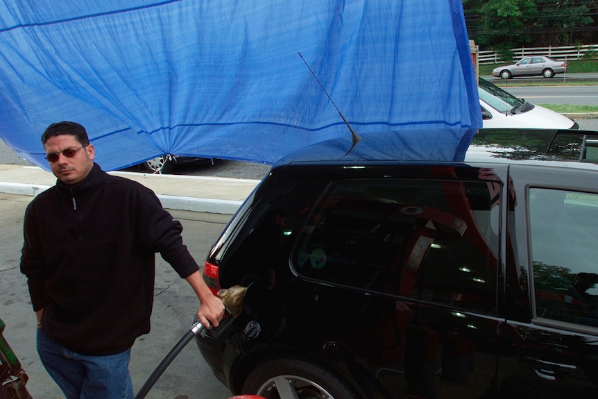 A man holds a petrol pump to his car in front of a blue tarpaulin