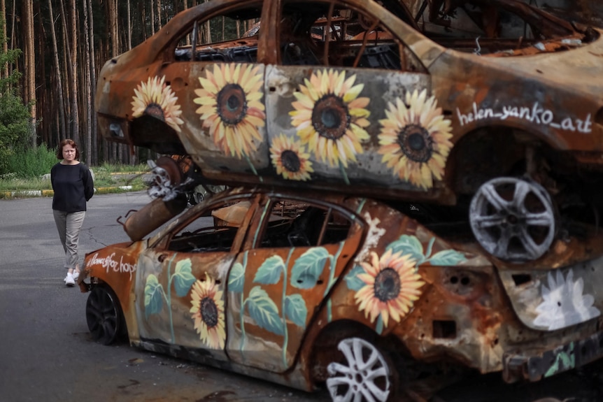 Due auto arrugginite bruciate accatastate l'una sull'altra con girasoli dipinti su un lato mentre una donna cammina a sinistra.