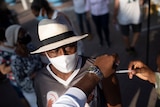 A dark-skinned man in white hat and face mask looks away as he is injected by white syringe