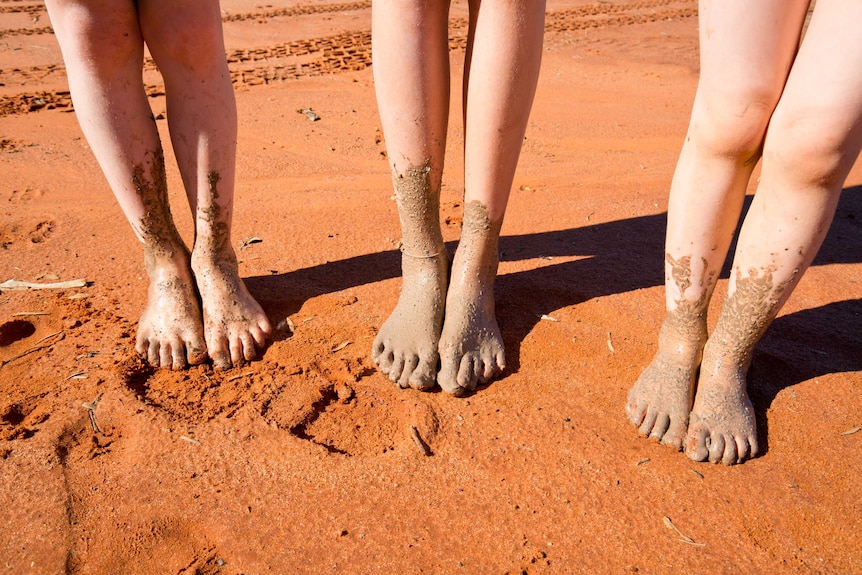 A close-up of three children's feet covered in mud as they stand in red sand.