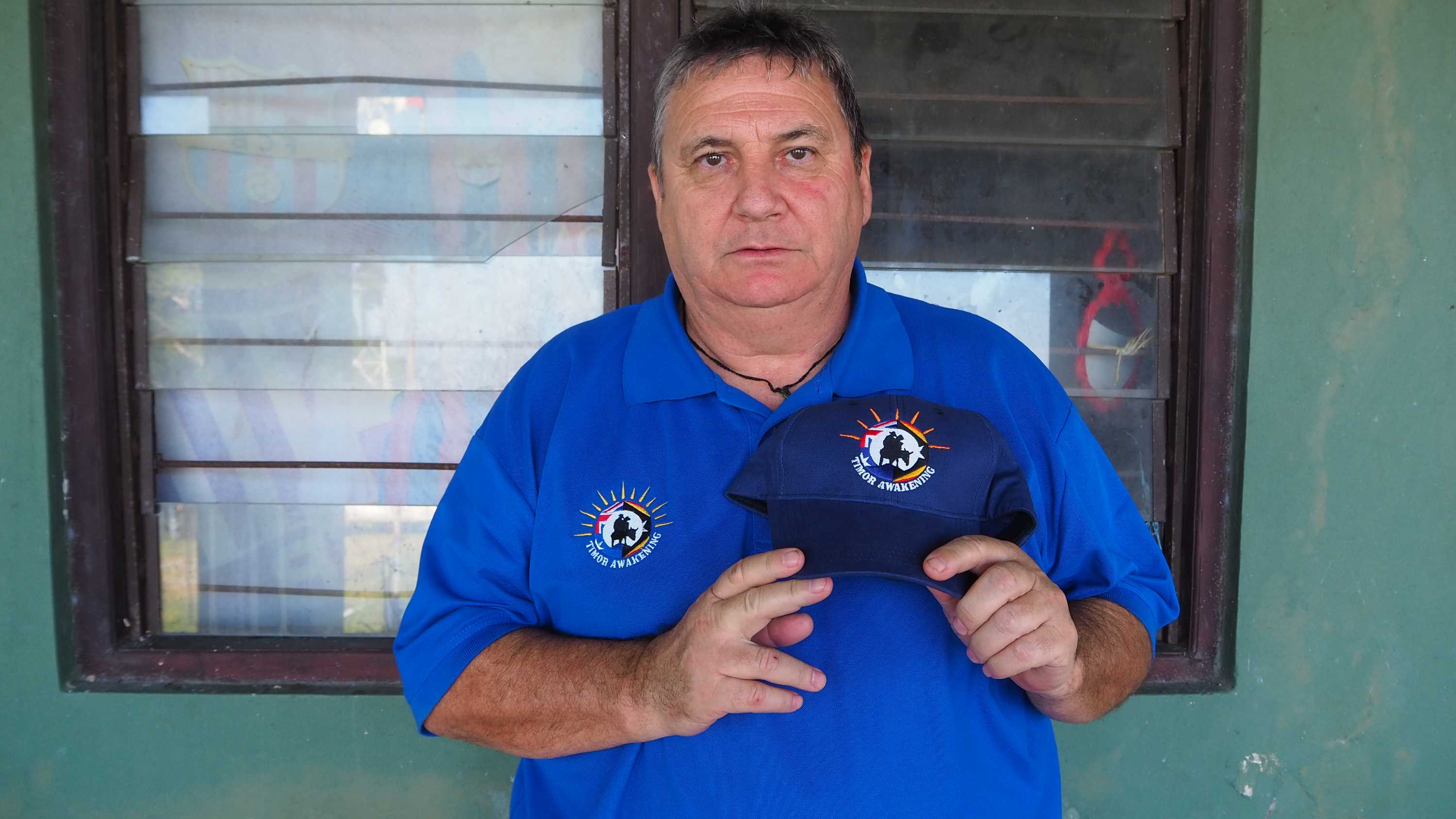 A man in a blue shirt holds up a matching blue cap next to his chest