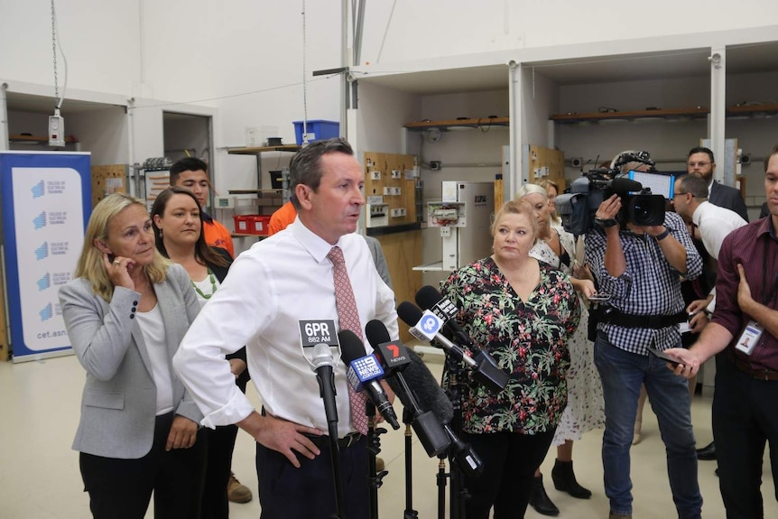 Mark McGowan with Sue Ellery standing in front of microphones in an apprentice workshop, taking questions from media.