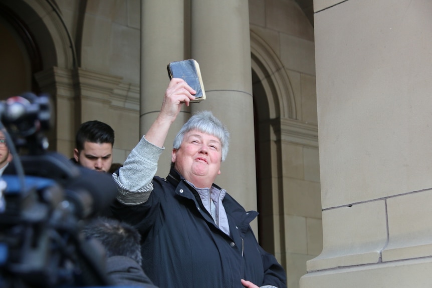 A woman with short grey hair holds a bible to the sky and smiles.