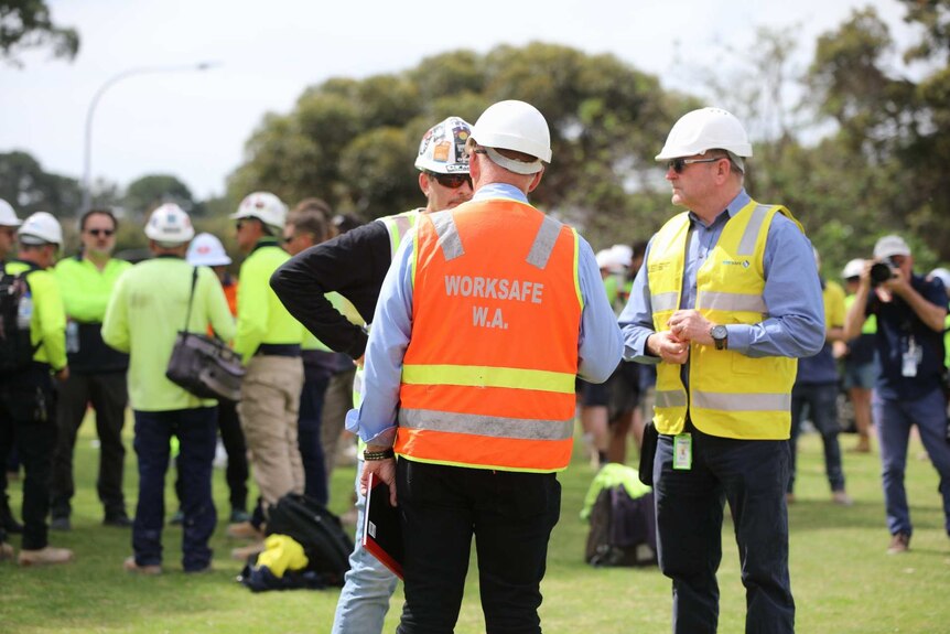 A man in an orange Worksafe WA hi-vis vest stands with has back turned to the camera talking to two workers.