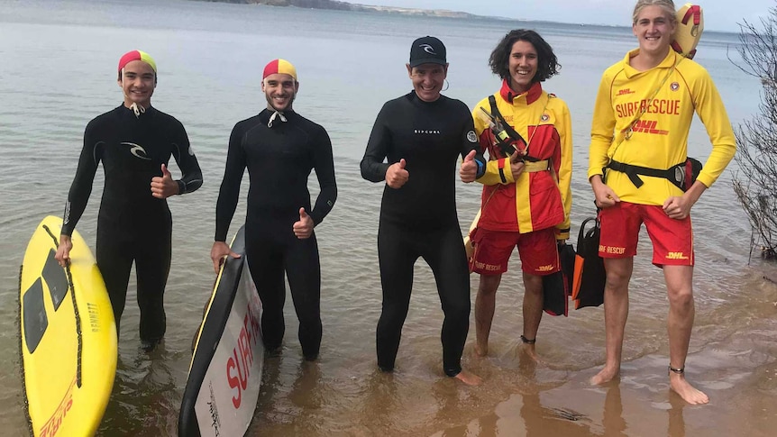 Three people in wet suits and two in surf lifesaving gear give the thumbs up as they stand together in the shallows at beach.
