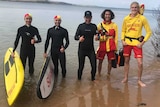 Three people in wet suits and two in surf lifesaving gear give the thumbs up as they stand together in the shallows at beach.