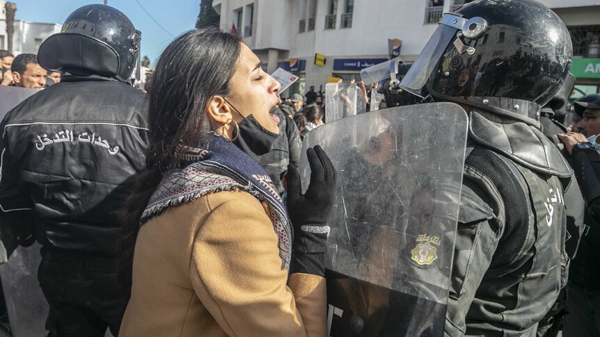 a woman in animated conversation with a riot police officer, who holds a shield between them
