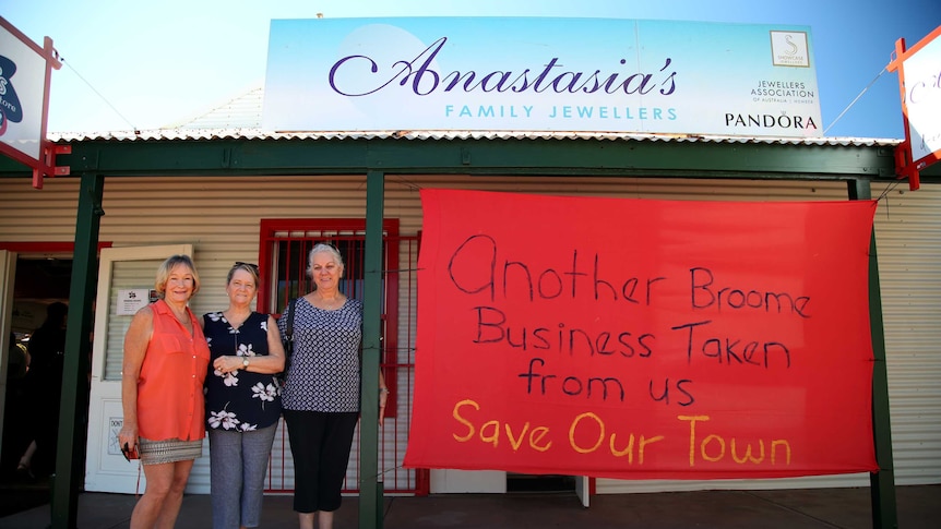 Three women stand on the verandah of a corrugated iron shop with the shop sign on top and a banner attached to the veranda.