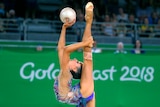A rhythmic gymnast holds her leg behind her back completely extended straight, and holds a ball in another hand.