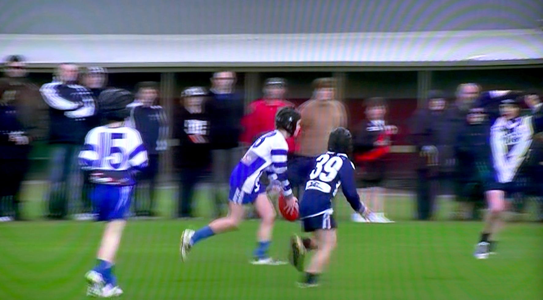 A still from video of boys playing Australian Rules football. One boy is running about to kick the ball.