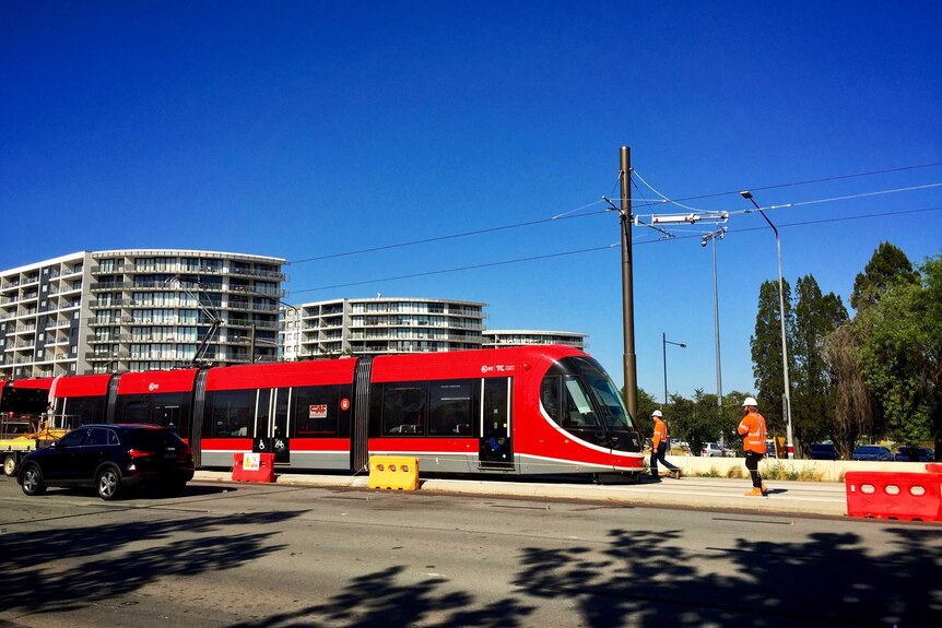 A bright red light rail tram sits idle on the tracks in Canberra with workers nearby.